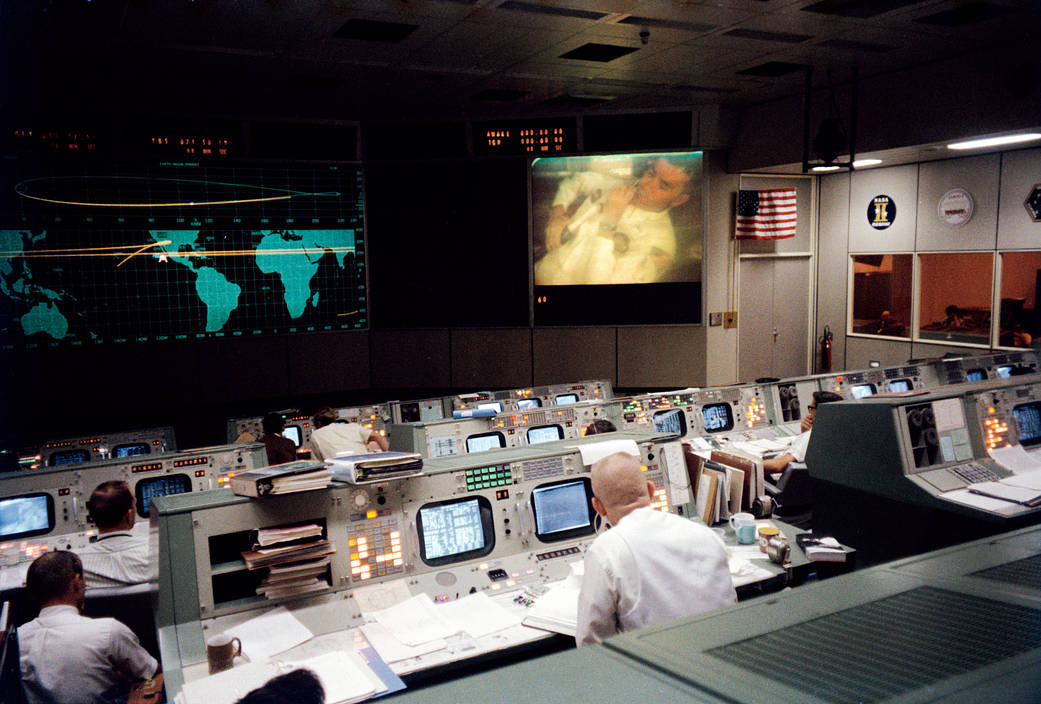 Mission Control room with flight directors facing monitors and large screens on walls showing view inside lunar module