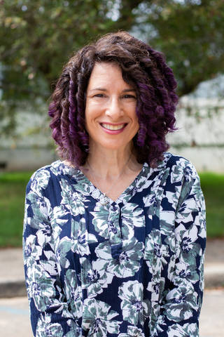Shari Miller, a white woman with shoulder-length ringlets of brown and purple hair, smiles at the camera in a portrait. She wears a blue and white floral blouse and silver necklace. Trees and asphalt are blurry in the background.