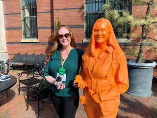 Wendy Bohon, a woman with long red hair, smiles and stands next to a bright orange statue of herself. She wears a green blouse and dark pants, and the red brick Smithsonian Castle walls are behind her.