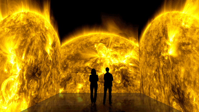 A moving GIF showing the black silhouettes of two people standing in front of a massive screen that stretches across three walls to their left, right, and in front of them. On the screen is projected a looping, swirling video of the Sun's surface, which is deep yellow and has bright yellow loops and dark yellow mottling on its surface.