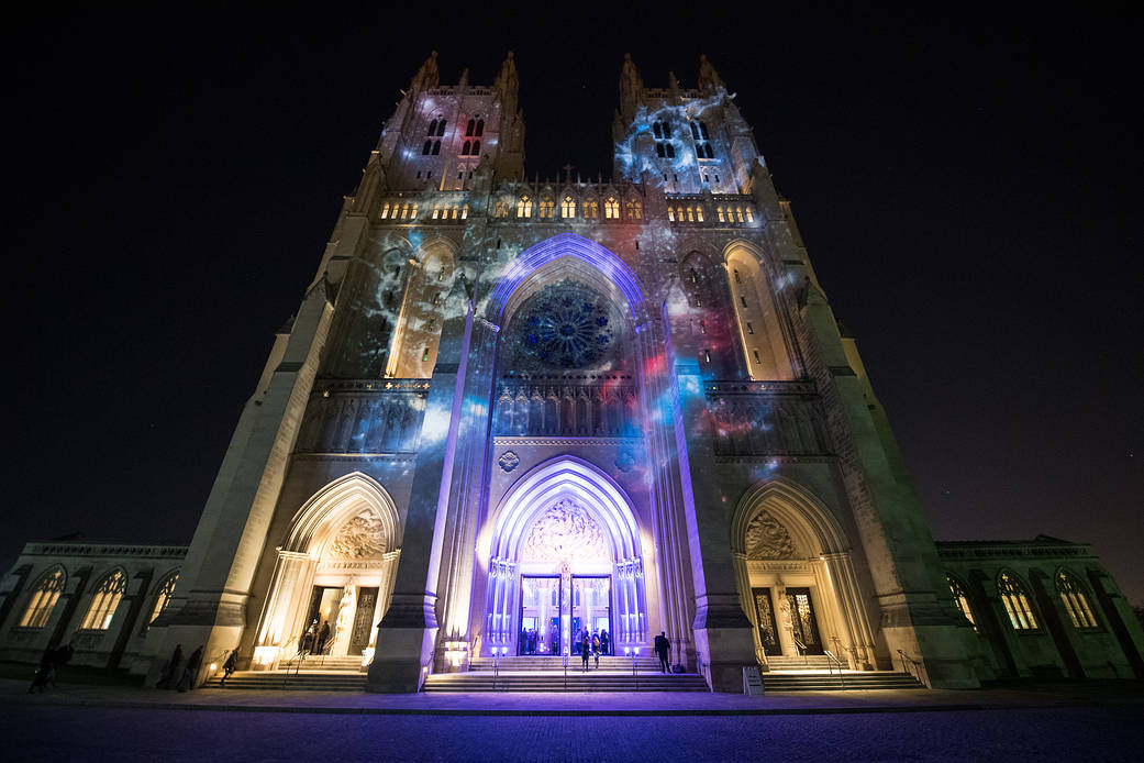 The facade of the Washington National Cathedral at nightfall with celestial galaxy projected on bricks in brilliant reds & blues