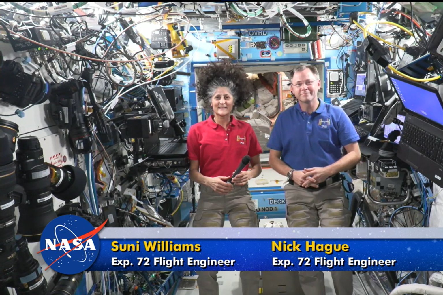 A screen capture of a STEM downlinks stream. Two astronauts are in the center of the image. The left female astronaut is wearing red, and the right male astronaut is wearing blue. There is a digital graphic near the bottom of the screen showing the NASA meatball, their names, and roles.