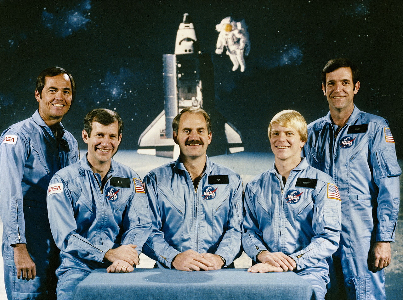 Five crew members in blue jumpsuits pose for portrait wit shuttle and man in spacesuit in the back drop