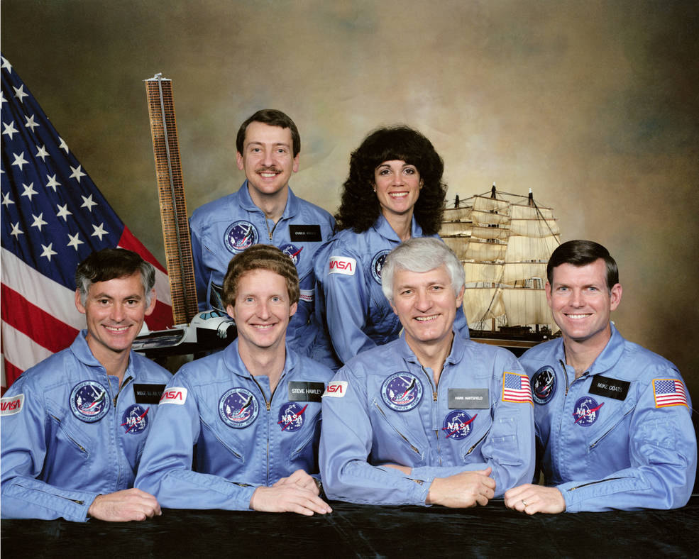 Six crew members pose in blue jumpsuits with model ship and US flag