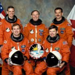 STS-74 crew members wearing their orange launch and entry suits with both the American and Canadian flags in the background.