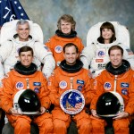 STS-76 crew wearing orange launch and entry suits and extravehicular mobility unit (EMU) suits. The American flag and Spirit of 76 flag are in the background.