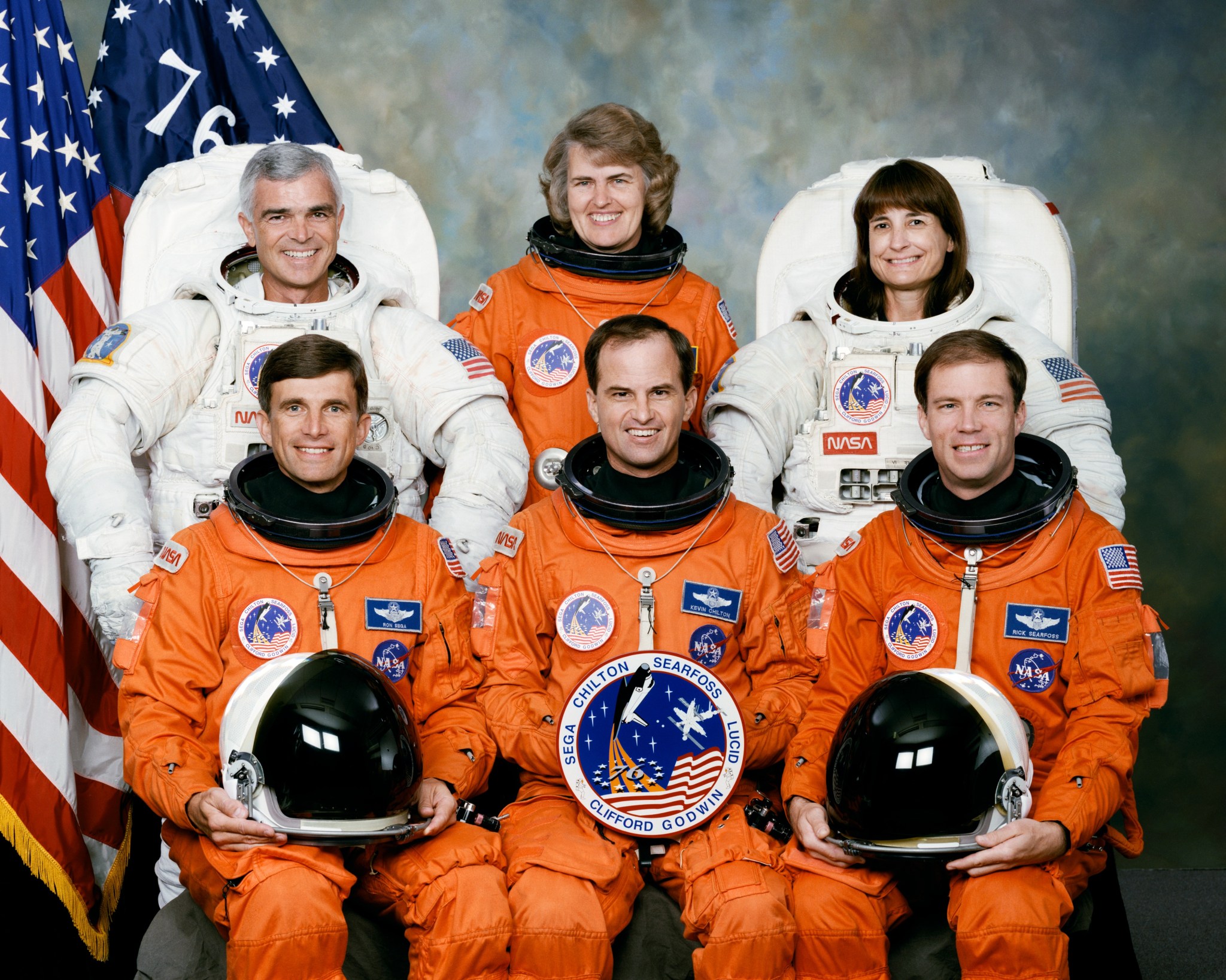STS-76 crew wearing orange launch and entry suits and extravehicular mobility unit (EMU) suits. The American flag and Spirit of 76 flag are in the background.