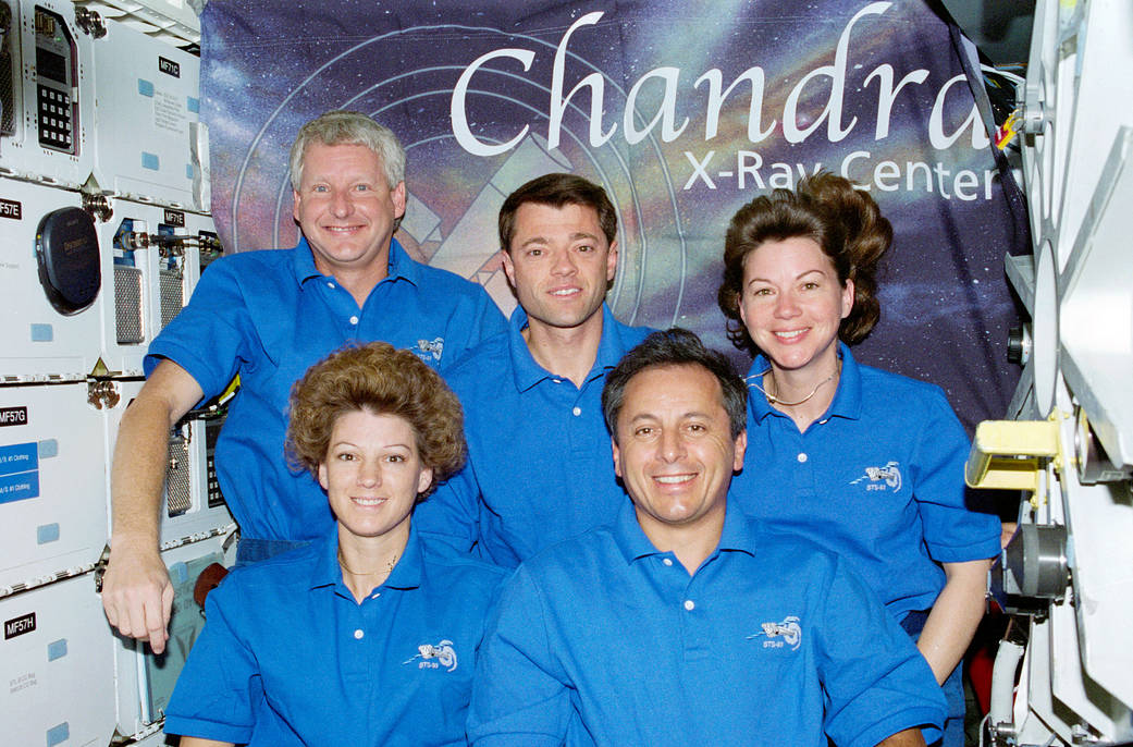 In this July 1999 photograph, five astronauts pose for a traditional inflight crew portrait with a large poster depicting the Ch