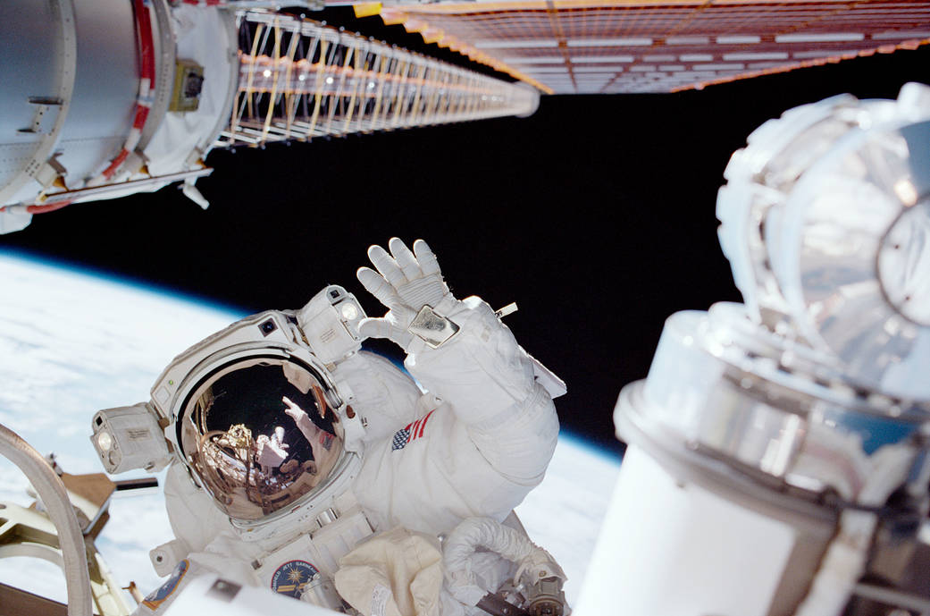Astronaut waves during spacewalk with space station solar array and Earth in background
