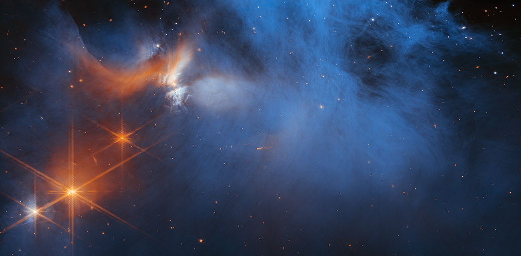 A black background filled with galaxies in shades of a red, orange and blue. In the foreground are blue smokey wisps. On the left top, the wisps are orange and white. Four bright points of light, three orange and one a white-orange mix at the bottom left.