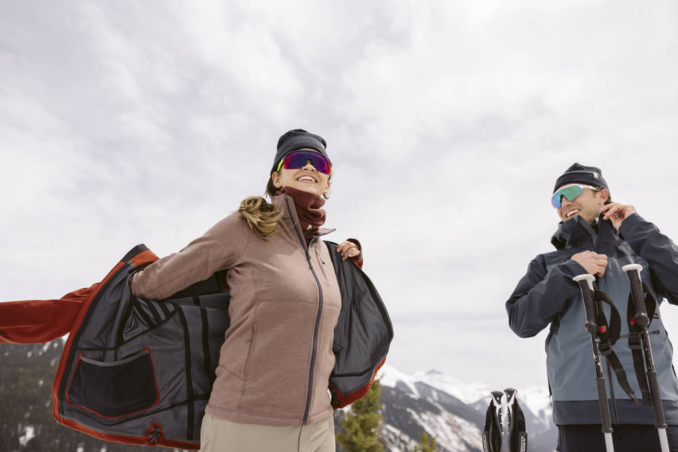 Man and woman in winter weather with ski gear and the woman in putting on a winter coat.