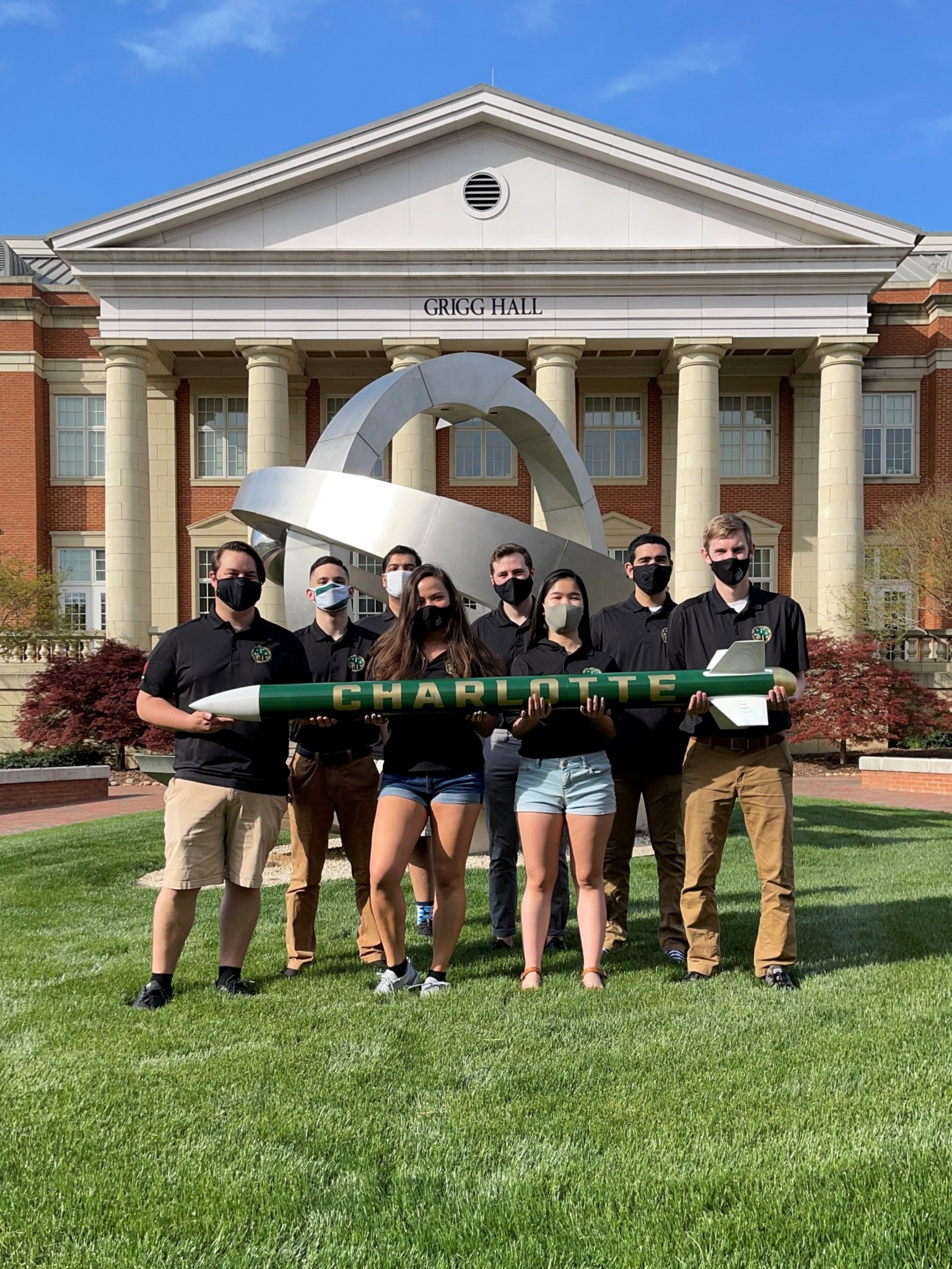 8 students from University of North Carolina standing in front of Grigg Hall holding "Charlotte" rocket