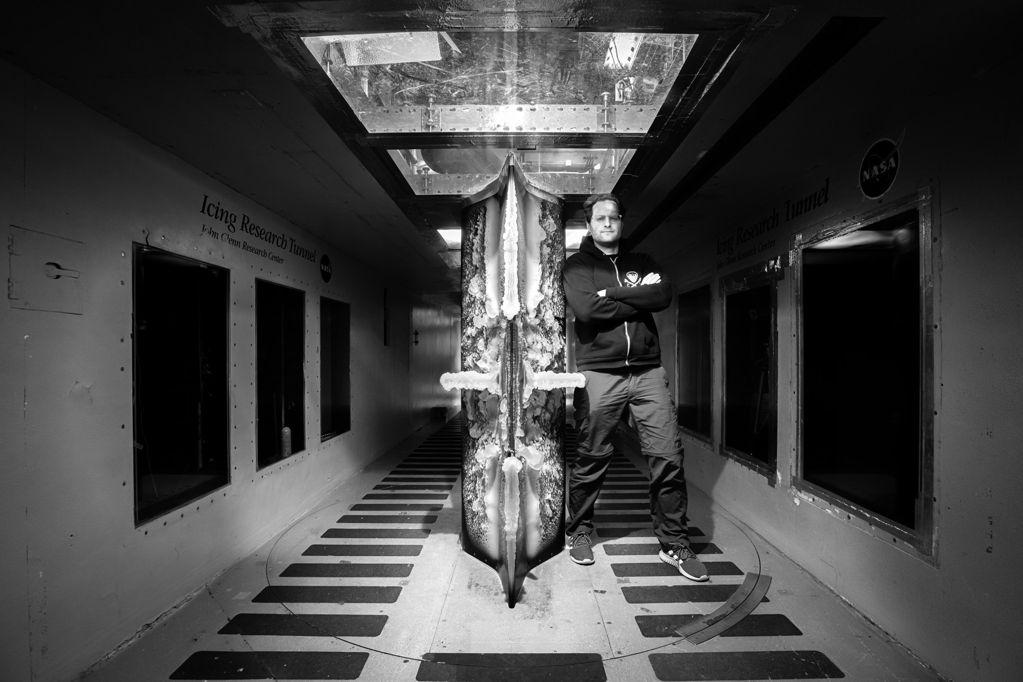 An airframe model is mounted vertically inside the Icing research tunnel. The test section of the tunnel is an enclosed corridor with large, square observation windows lining the walls.  The airframe has layers of ice covering its surface. A NASA researcher stands next the airframe. He is facing the camera with his arms crossed and wears glasses, jeans, and a hoodie. The photo is a black and white image. 