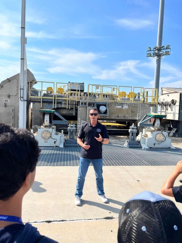 A man in a black tshirt, jeans and dark sunglasses stands in front of a rocket launch pad, a 12 foot tall metal and concrete structure with yellow railings
