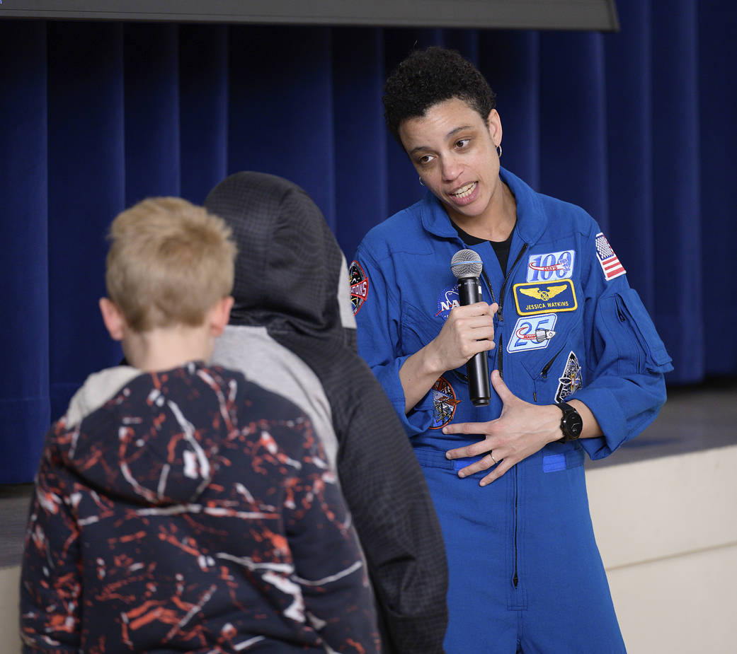 Two elementary school children facing away from the camera talking to astronaut Jessica Watkins, wearing a blue jump suit. Watkins is holding a microphone and her head is tilted towards the children.