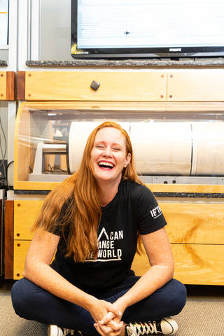 Wendy Bohon, a woman with long red hair, sits on the floor in front of a large piece of equipment and laughs. She wears a black tee, jeans, and black and white sneakers. The equipment looks like a large wooden cabinet with a spool visible through glass.