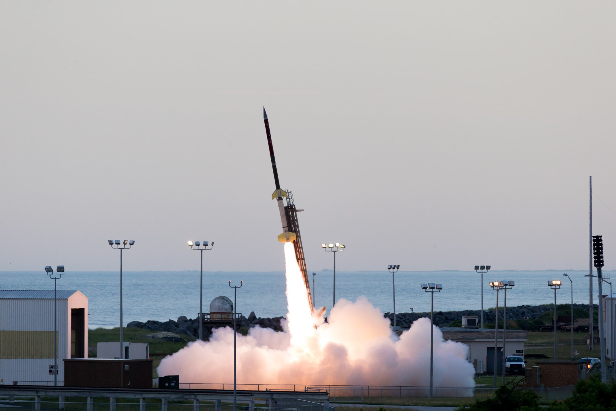 A sounding rocket seconds in mid-launch off the launch pad with a bright white plume underneath. In the background is the Atlantic Ocean with a pale grey sky with no clouds.