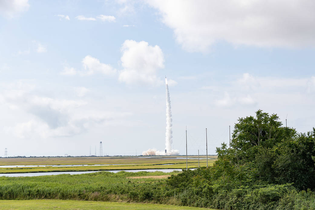 Minotaur I rocket launching across blue cloud sky with green grass in the foreground.