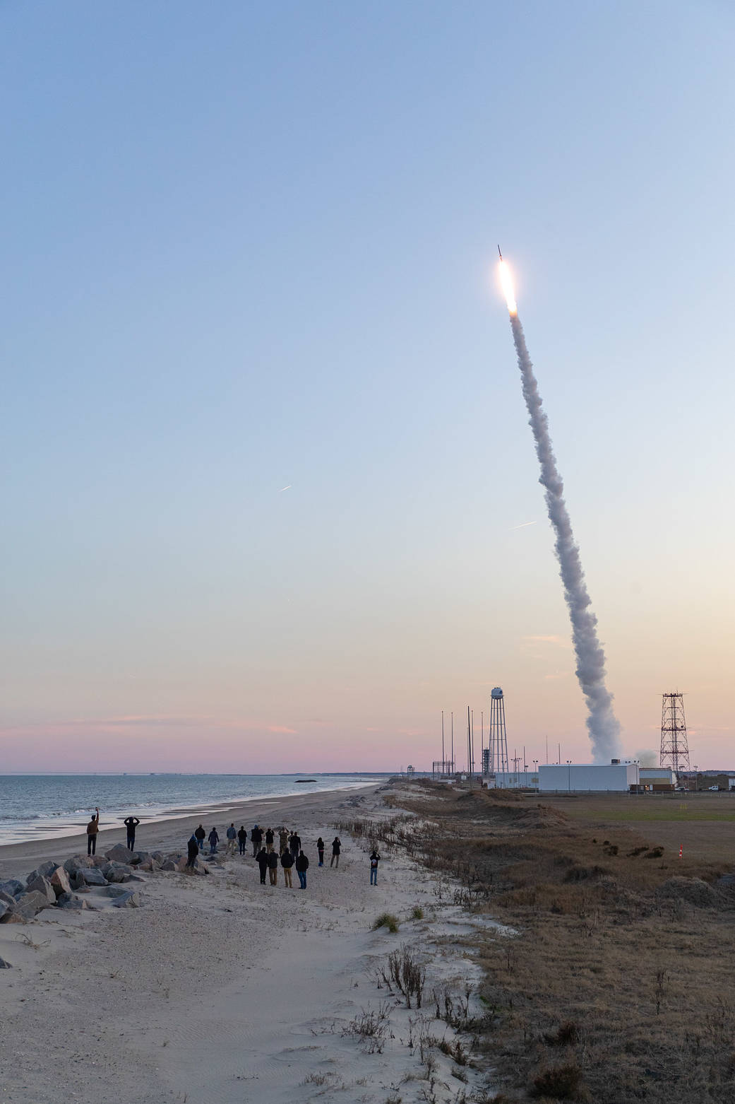 landscape photo with a small crowd of people in the foreground standing on a beach while watching the sounding rocket launch in the background against a dusky sky.
