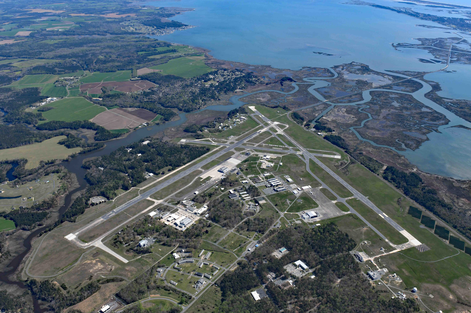 An aerial photo of Wallops Flight Facility, showing the center's airstrips, buildings, and parking lots with the Chesapeake Bay shining blue across the upper right corner of the image. The center has two perpendicular airstrips connected by a diagonal taxi strip, and these frame the buildings and parking lots in the center. Past the edges of the airstrip are trees, shoreline areas and dark, snaking water.