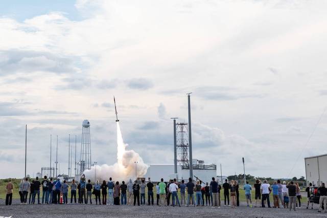 In the foreground, a line of people stand at a safe distance away from a sounding rocket launching in the background. The rocket is just off the pad with a plume of fire and smoke underneath.