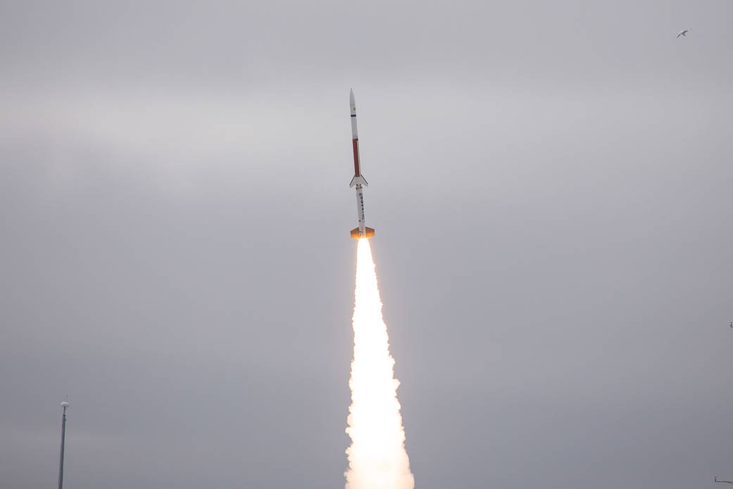 A sounding rocket seconds after launch with a bright white plume underneath. In the background is a pale grey overcast sky.