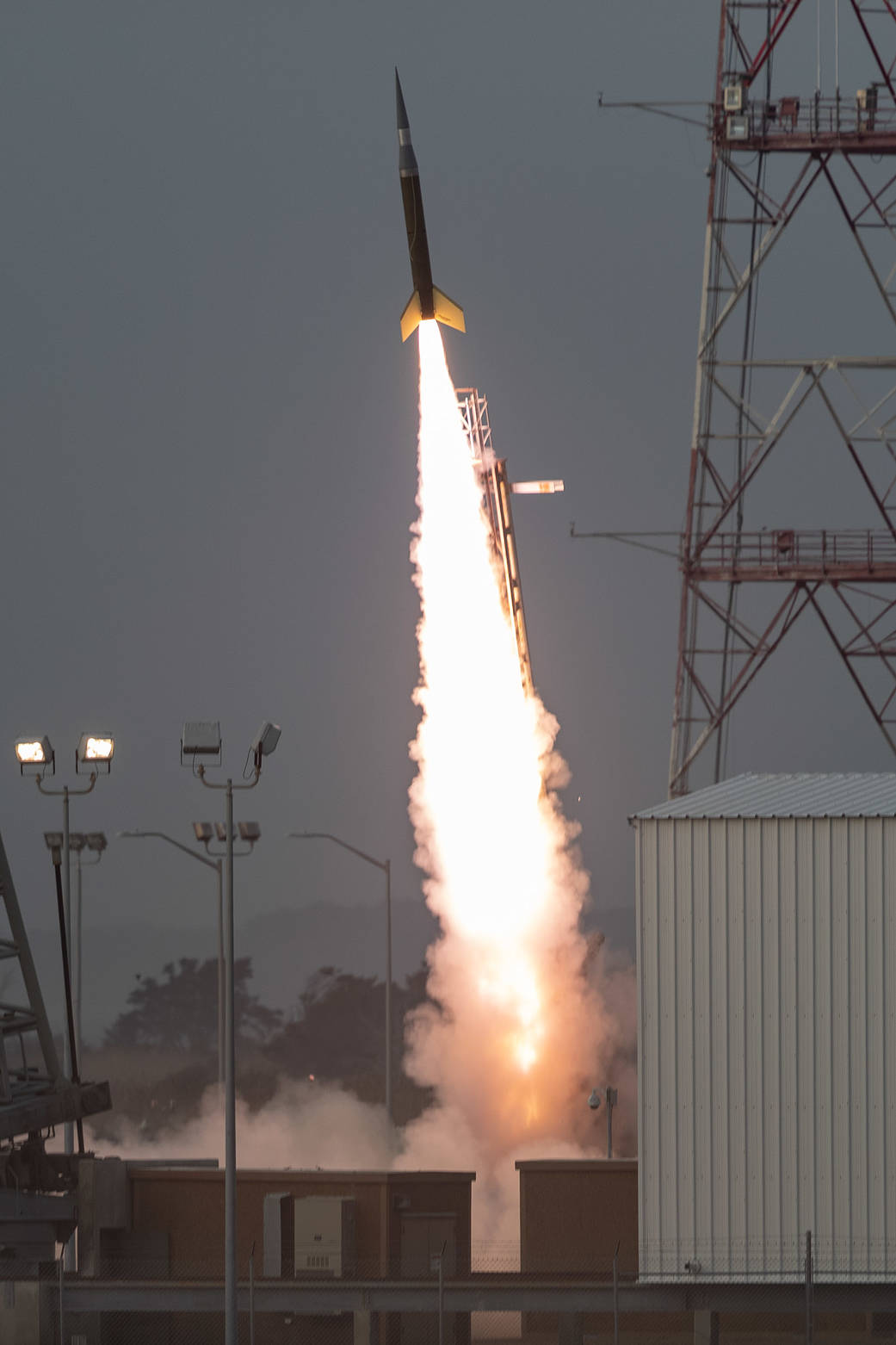 A small sounding rocket launching into the air with a bright white smoke plug trailing underneath. The rocket body is dark colored with yellow fins on the bottom. The sky is a pale blue color.