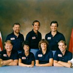 Crew poses in matching NASA shirts for crew portrait in front of US and Canadian flag