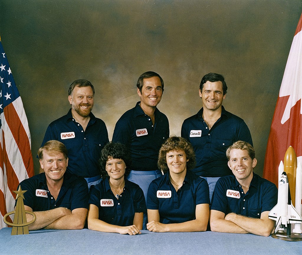 Crew poses in matching NASA shirts for crew portrait in front of US and Canadian flag