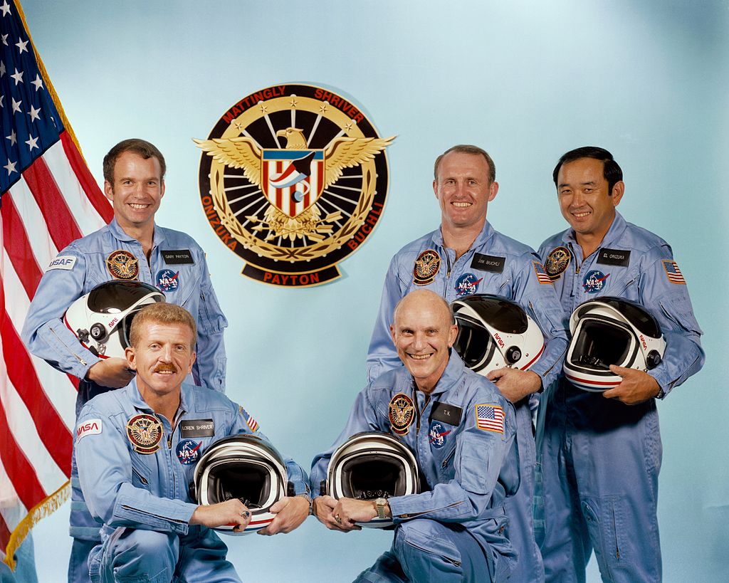 Five men smile for their group picture. They are wearing space flight suits. They are holding space suite helmets in their hands. 