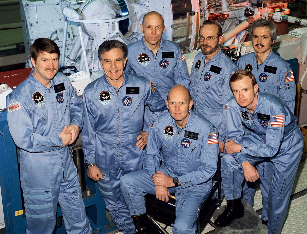 Seven men stand and smile for their group photo. They are wearing space flight suites with NASA emblems on them. 