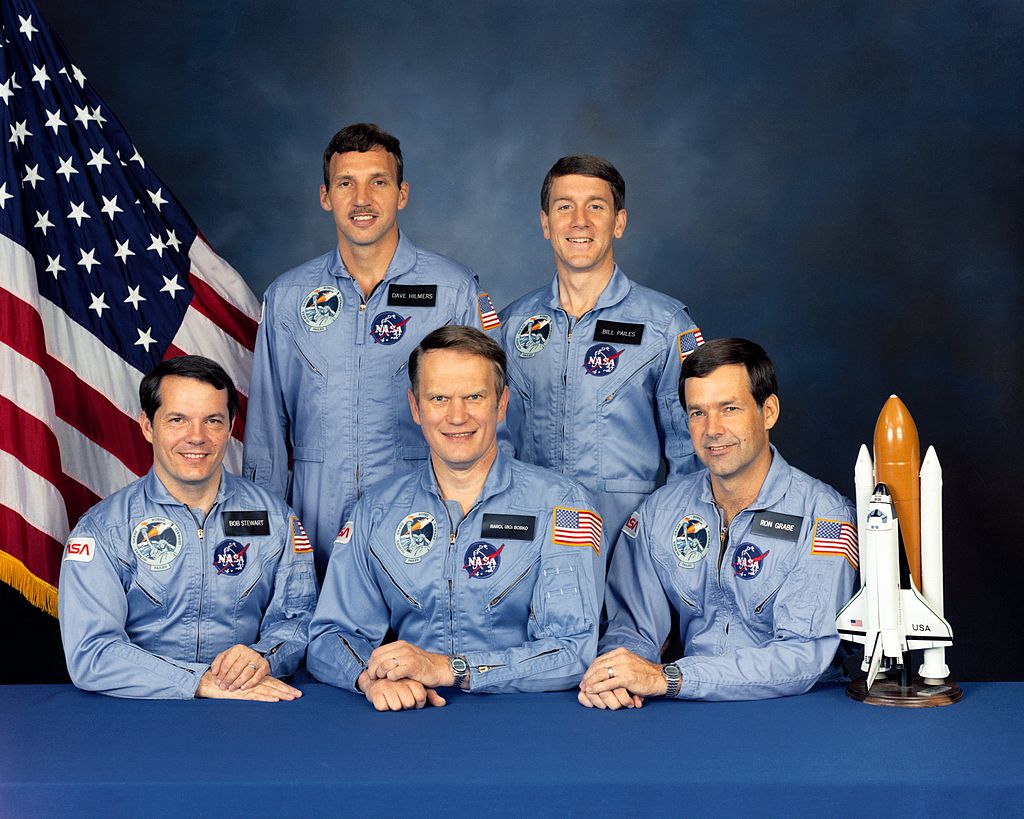 Five astronauts pose for their group portrait. They are all wearing space flight suites. There is a small model rocket ship next to them.