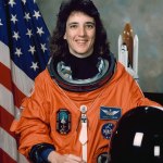 A woman smiles, wearing a spacesuit, and stands in front of the American flag. He has his helmet off and sitting on table before him.