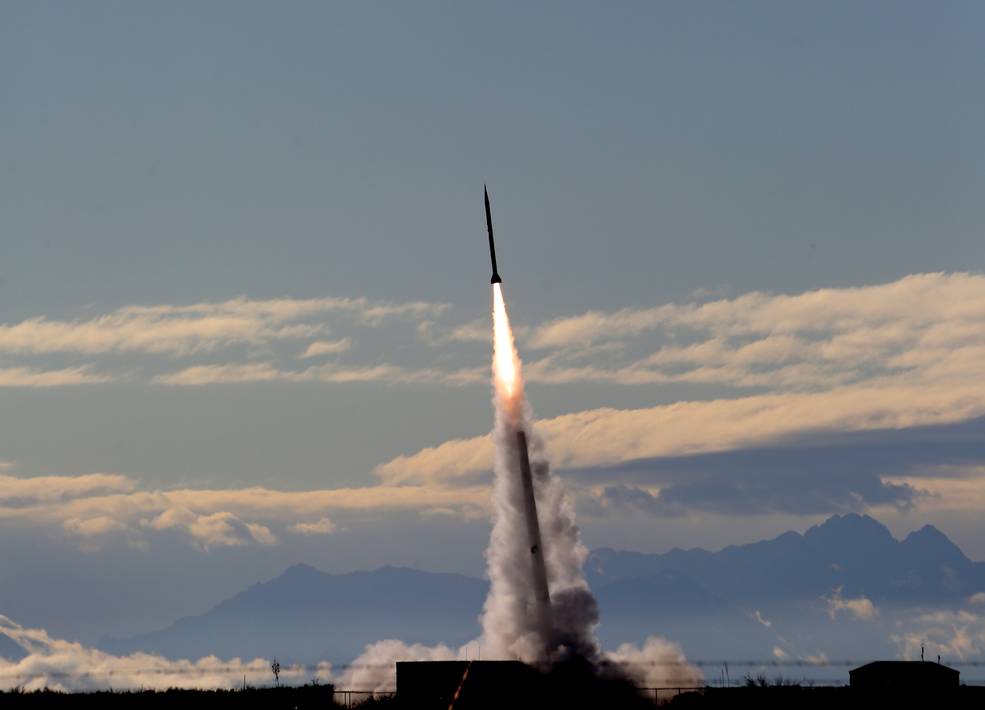 A suborbital launch vehicle taking off into a grey-blue sky with clouds hovering over mountains in the distance.