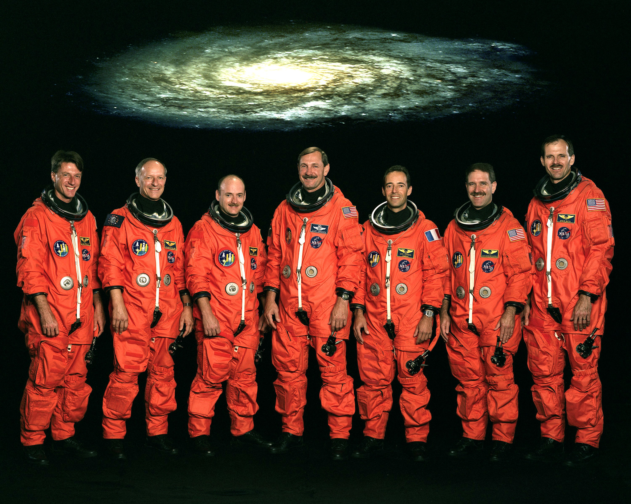 Seven men in flight suits, with no helmets, standing in front of a galaxy image