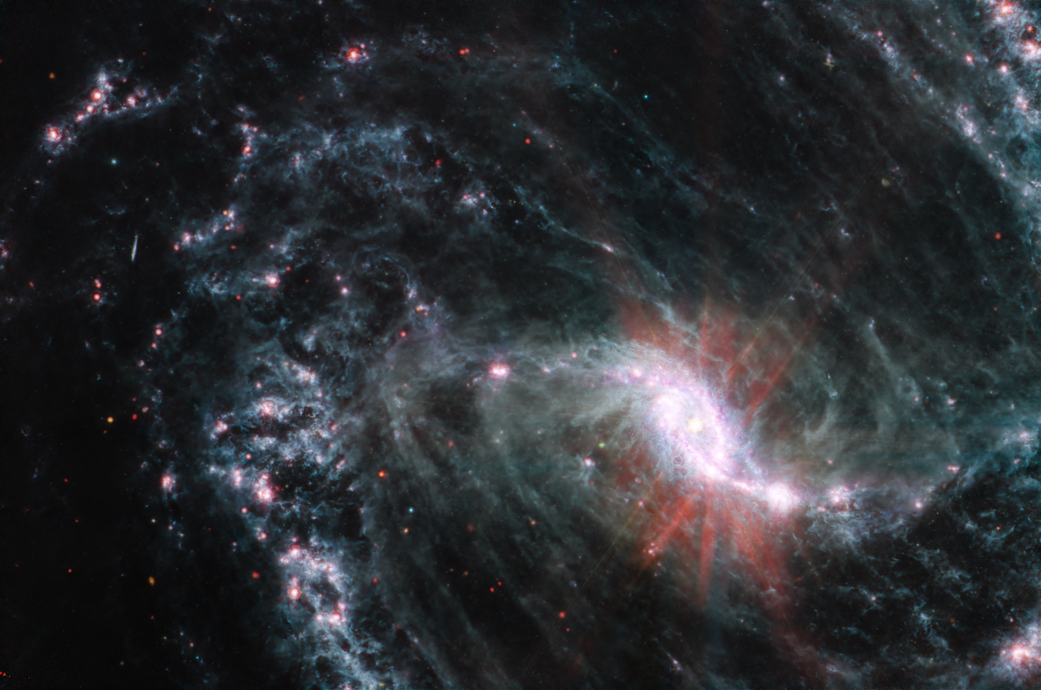 Close-up of a wispy spiral galaxy on a black background. It features a glowing, light pink core in an elongated oval shape. Red streaks appear to emanate out of the core.