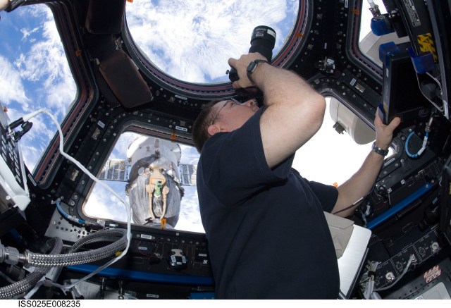 male astronaut holding a camera and pointing it out a window in the cupola of the space station while taking pictures of Earth