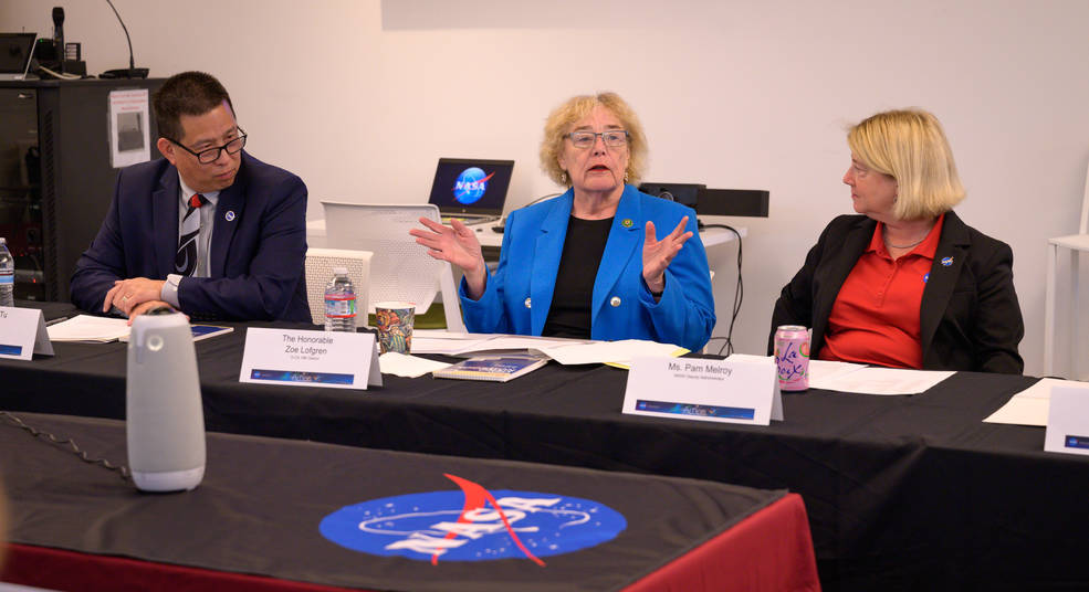 A man in a dark blue suit, woman in a royal blue blazer, and a woman in a red shirt and black blazer sit at a conference room table with a NASA logo visible.