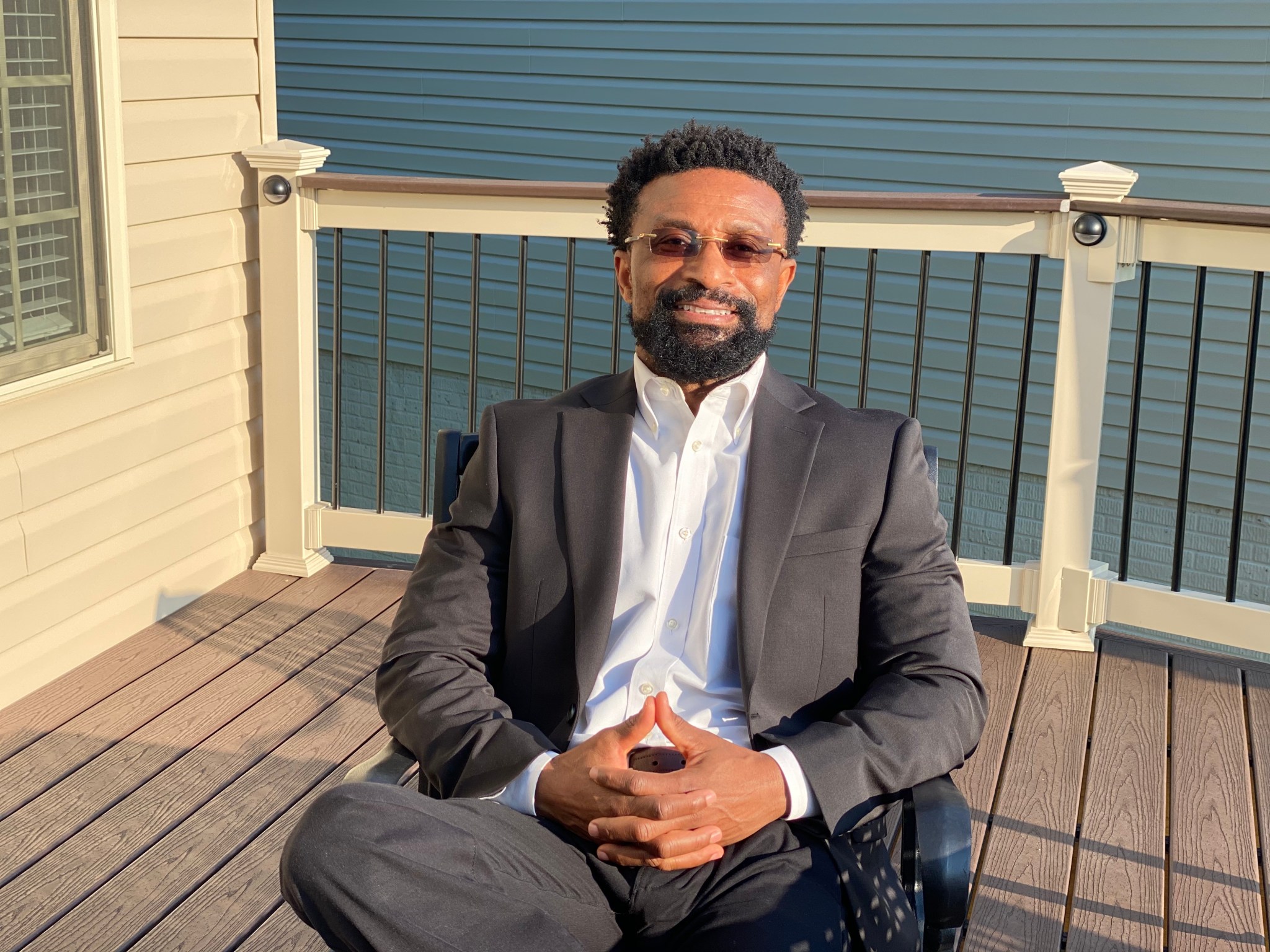 Moses Adoko, a man with short, curly black hair and beard, smiles and relaxes in a chair on a sunny deck. He wears a gray suit with white shirt, and small tinted glasses.