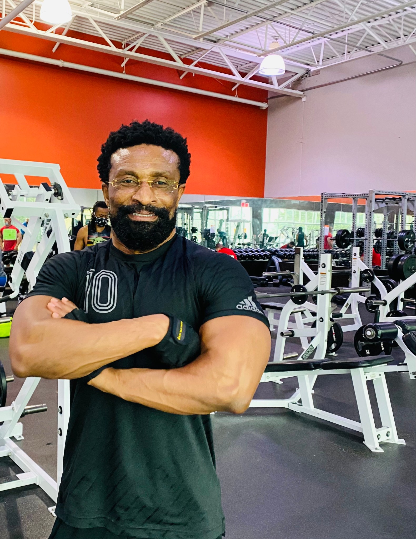 Moses Adoko, a man with short, curly black hair and beard, smiles and stands with arms crossed in a large gym. He wears a fitted black tee with the number 10, black fingerless gloves, and small rimless glasses. The gym wall is bright orange.