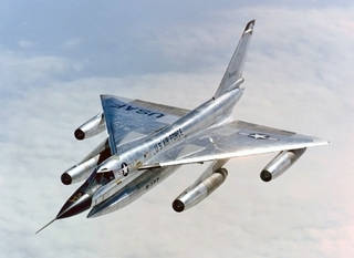 An all-silver supersonic nuclear bomber with four jet engines and a triangle-shaped wing flies high over some clouds.