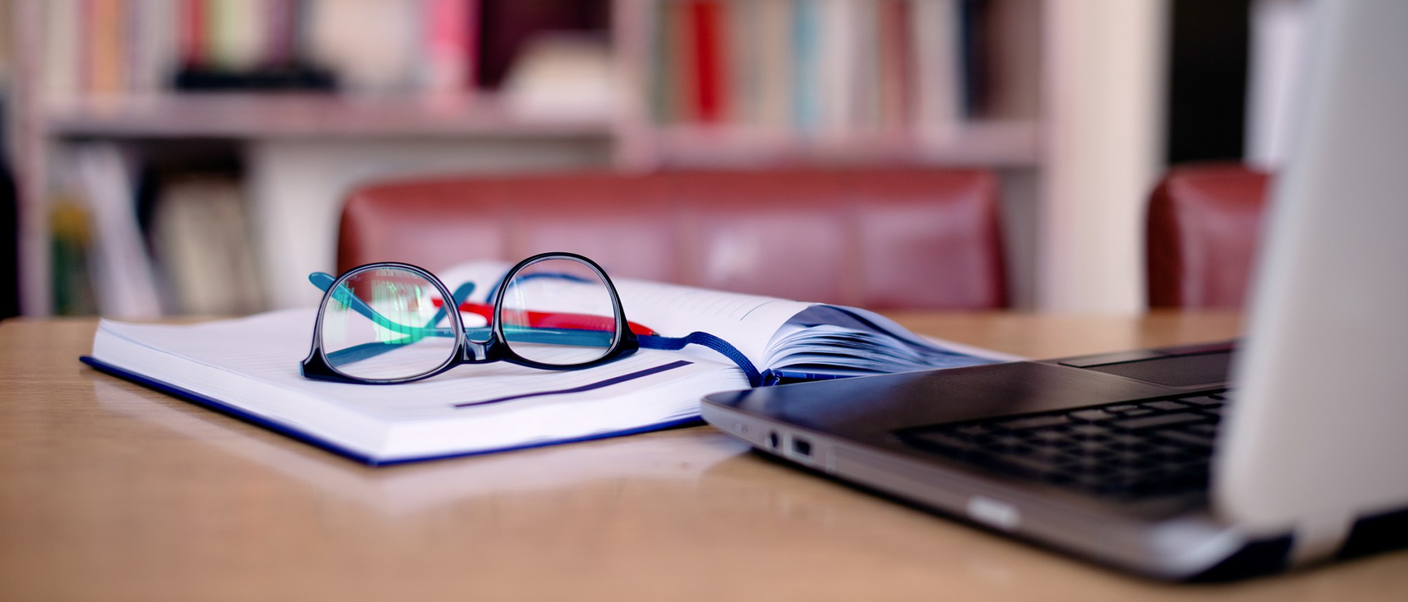 Image of a desk with a notebook with eyeglasses on top and a laptop.