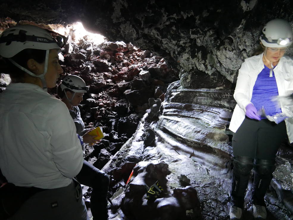 Three scientists, wearing helmets, stand in the foreground of this image, which is taken in a dark cave that's covered in boulders on the ground. An opening in the background of the image, like an open mouth, streams in a bit of daylight.