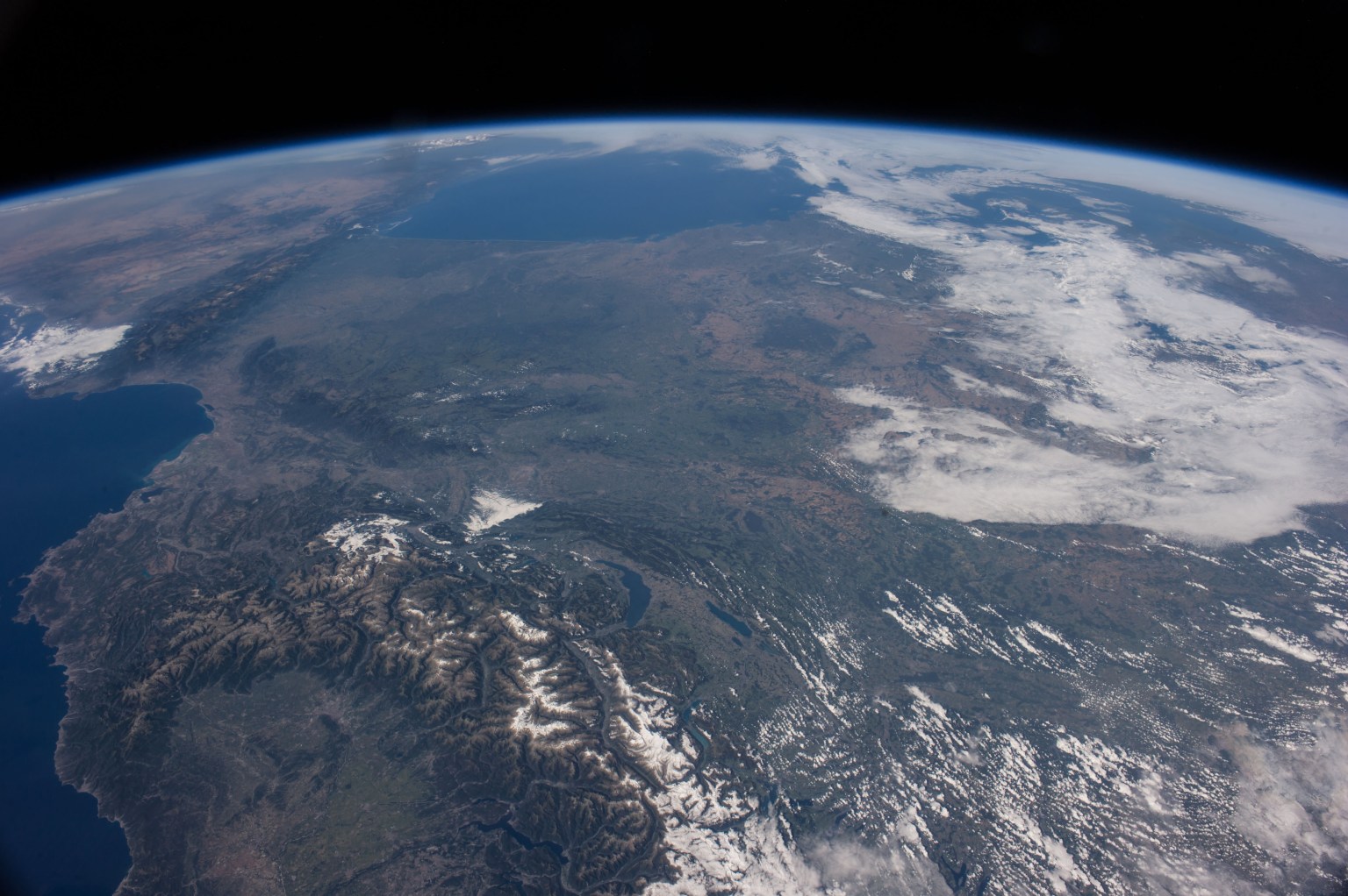A photograph of Earth taken from orbit, with green and brown land dominating much of the image and blue ocean visible near the top of the planet's curved edge. In the foreground, mountain ridges cover the land in branching patterns, with snow dusting the tops of some peaks. White clouds cover the upper right portion of Earth's surface.