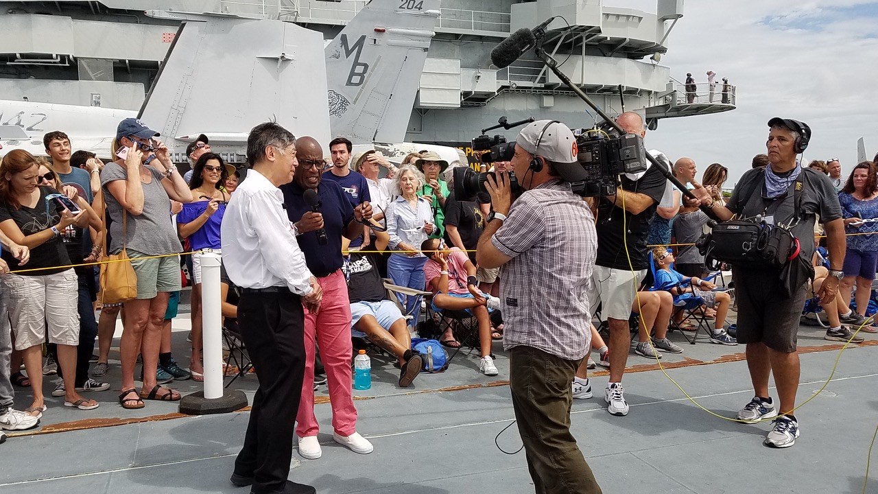 Dr. Gordon Chin, a man with graying dark hair and glasses, stands on the deck of an aircraft carrier being interviewed for a TV show. He wears a white shirt and black pants. Two men with a camera and a large microphone stand in front of him.