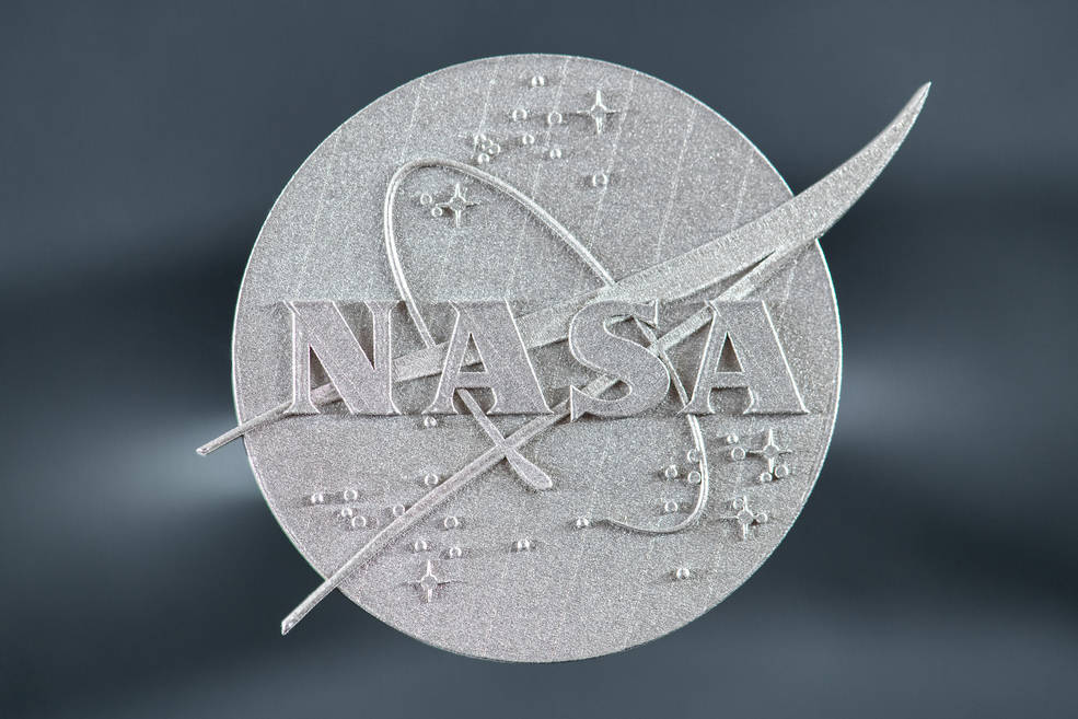 A close-up view of a 3D printed NASA logo is shown against a gray background.