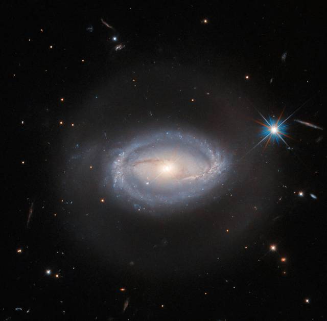 Spiral galaxy: 2 almost-straight arms coming from the left and right of the golden core that meet a starry, bluish ring around the edge. A faint light halo surrounds the galaxy. One bright star, a few small stars and galaxies all on a black background.