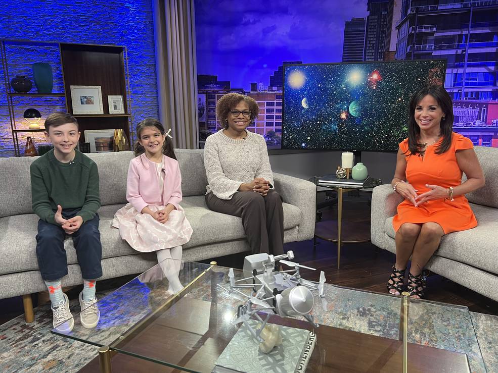 Two children and an adult woman sit on a couch. Another adult woman in an orange dress sits on an opposite couch in a television studio.