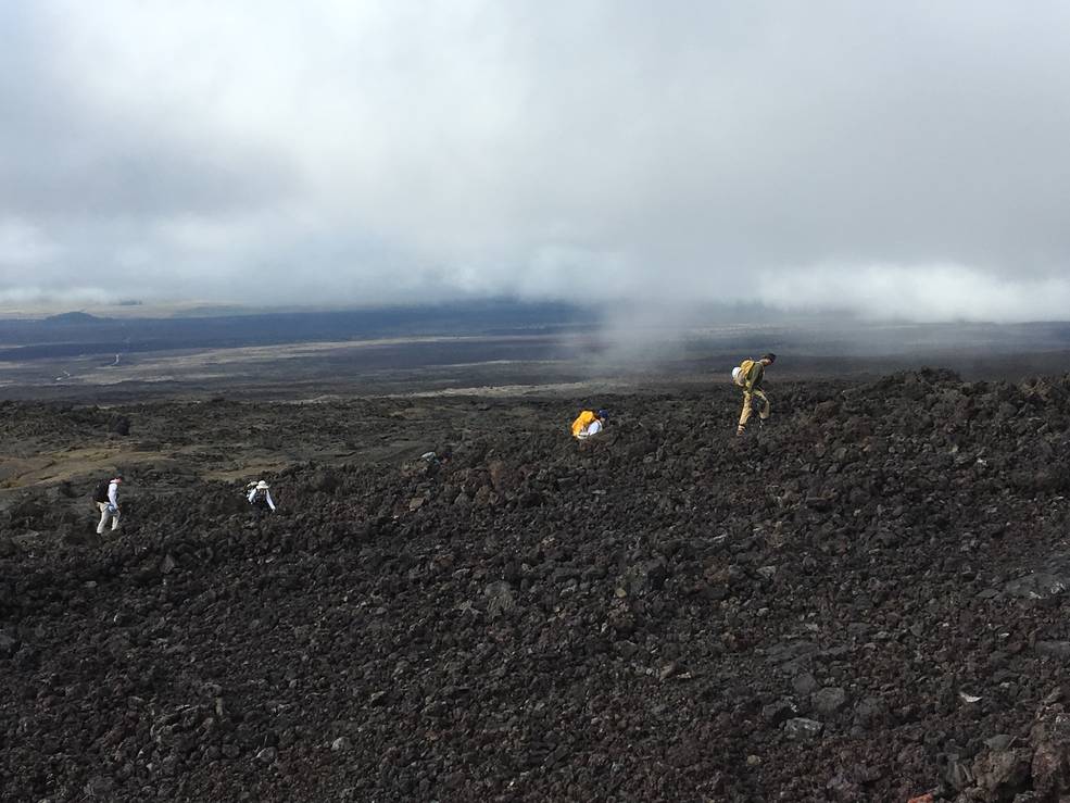 This images shows a large vista at a Hawaiian volcano. In the foreground are dark, jagged rocks. A few people with backpacks are walking. Smoky clouds hover overhead.