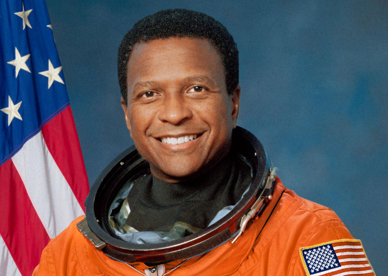Official Portrait of Astronaut Mike Anderson (STS-107) in his orange launch and landing suit with an American flag in the background.
